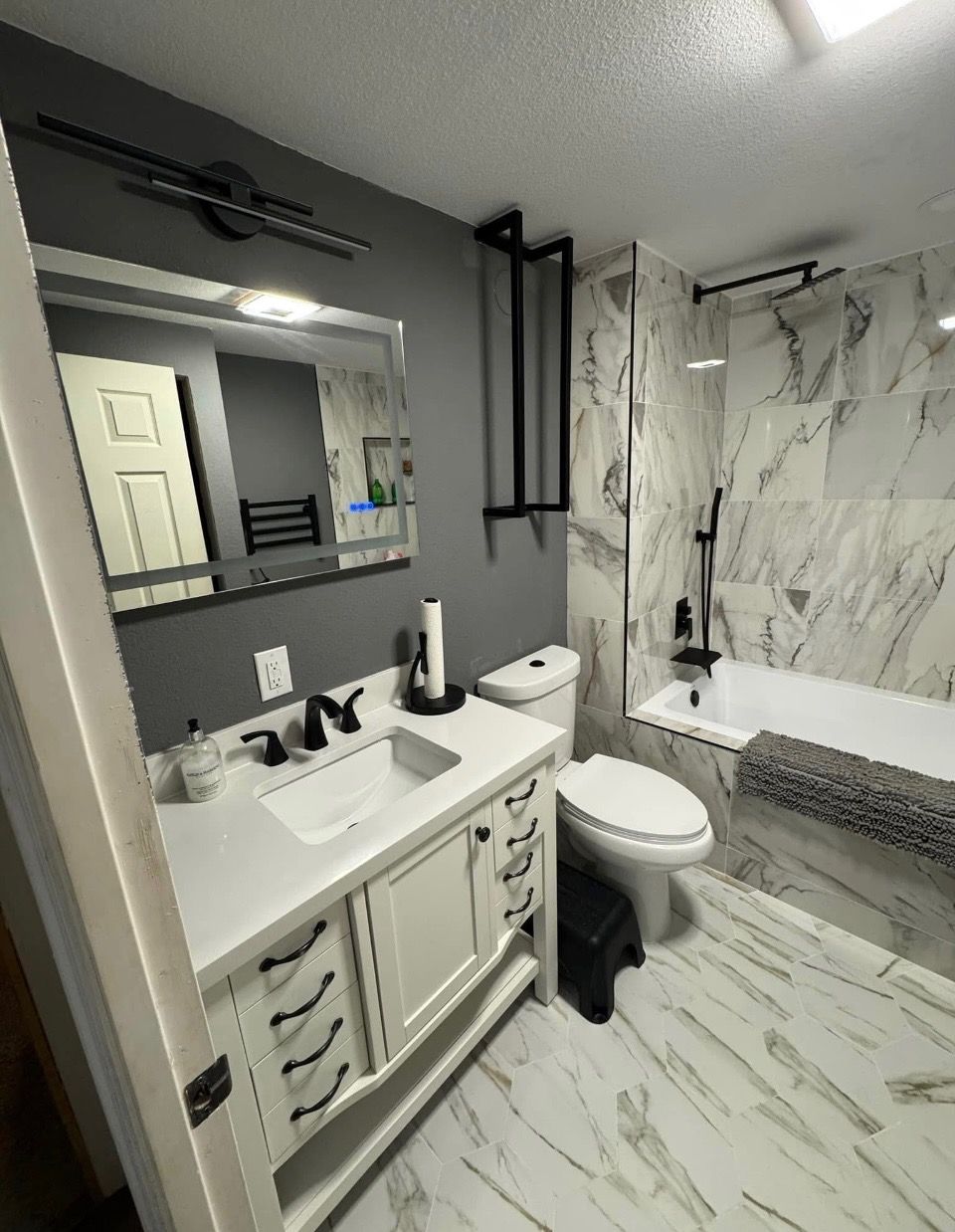 The same bathroom after remodeling, with a white vanity, quartz top, marble-look tile surround and hexagon marble floor