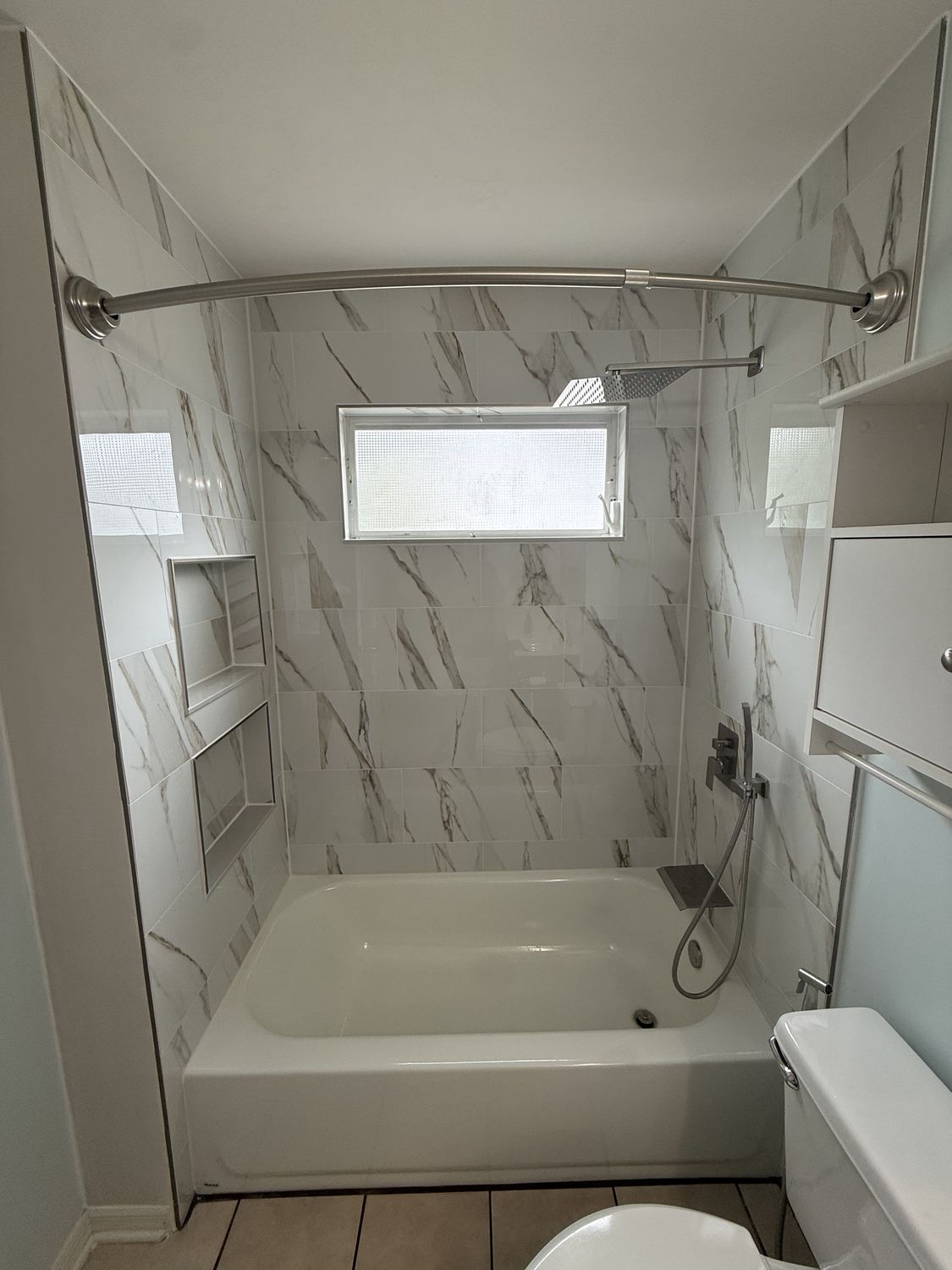 The same bathroom after remodeling, with large-format marble-look porcelain, a recessed niche and a curved shower rod