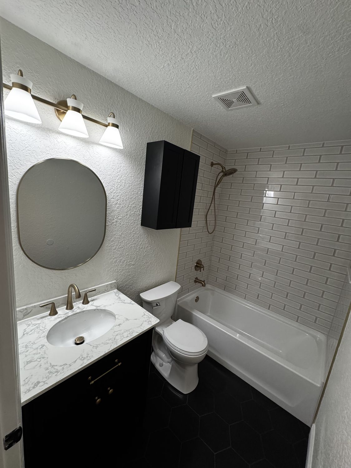 The same bathroom after remodeling, with a black vanity and marble-look top, oval mirror, brass lighting and a subway-tile tub surround