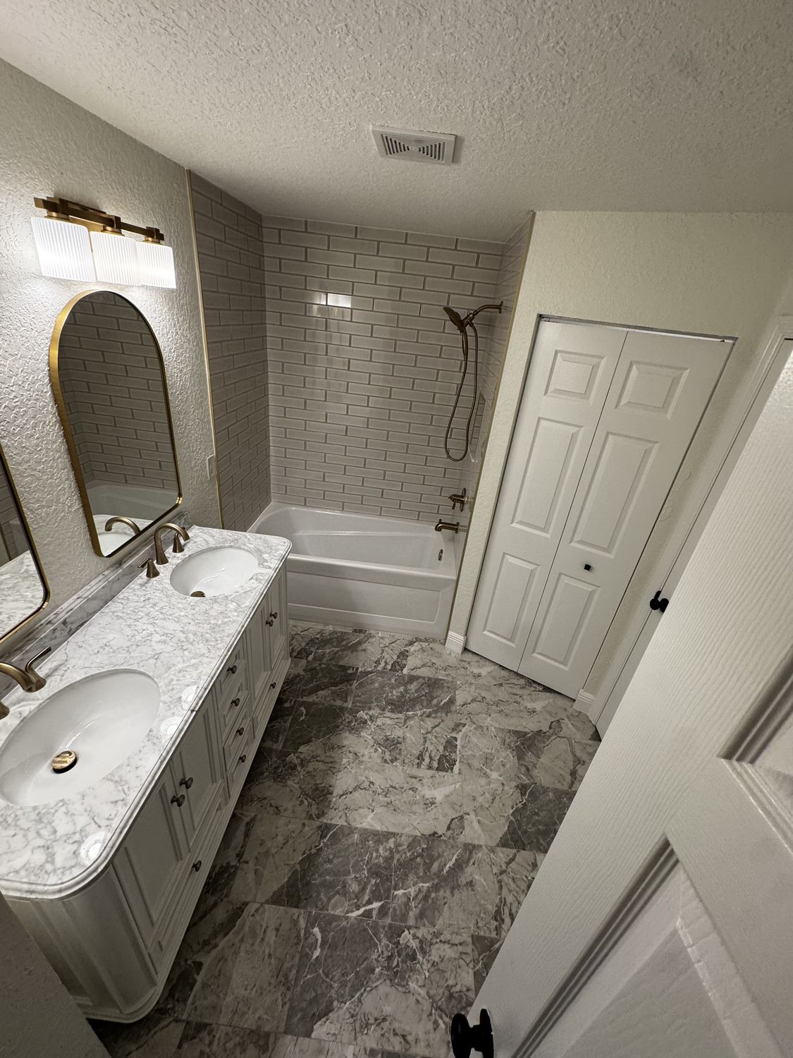 The same bathroom after remodeling, with a marble-topped double vanity, arched mirror, subway-tile tub surround and grey marble-look floor