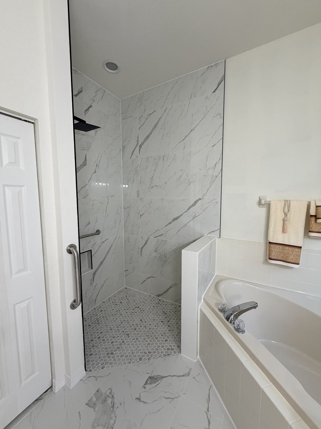 The same master bathroom after remodeling, finished floor to ceiling in marble-look porcelain with an open shower, pebble shower floor, matte black fixtures and a re-tiled tub deck