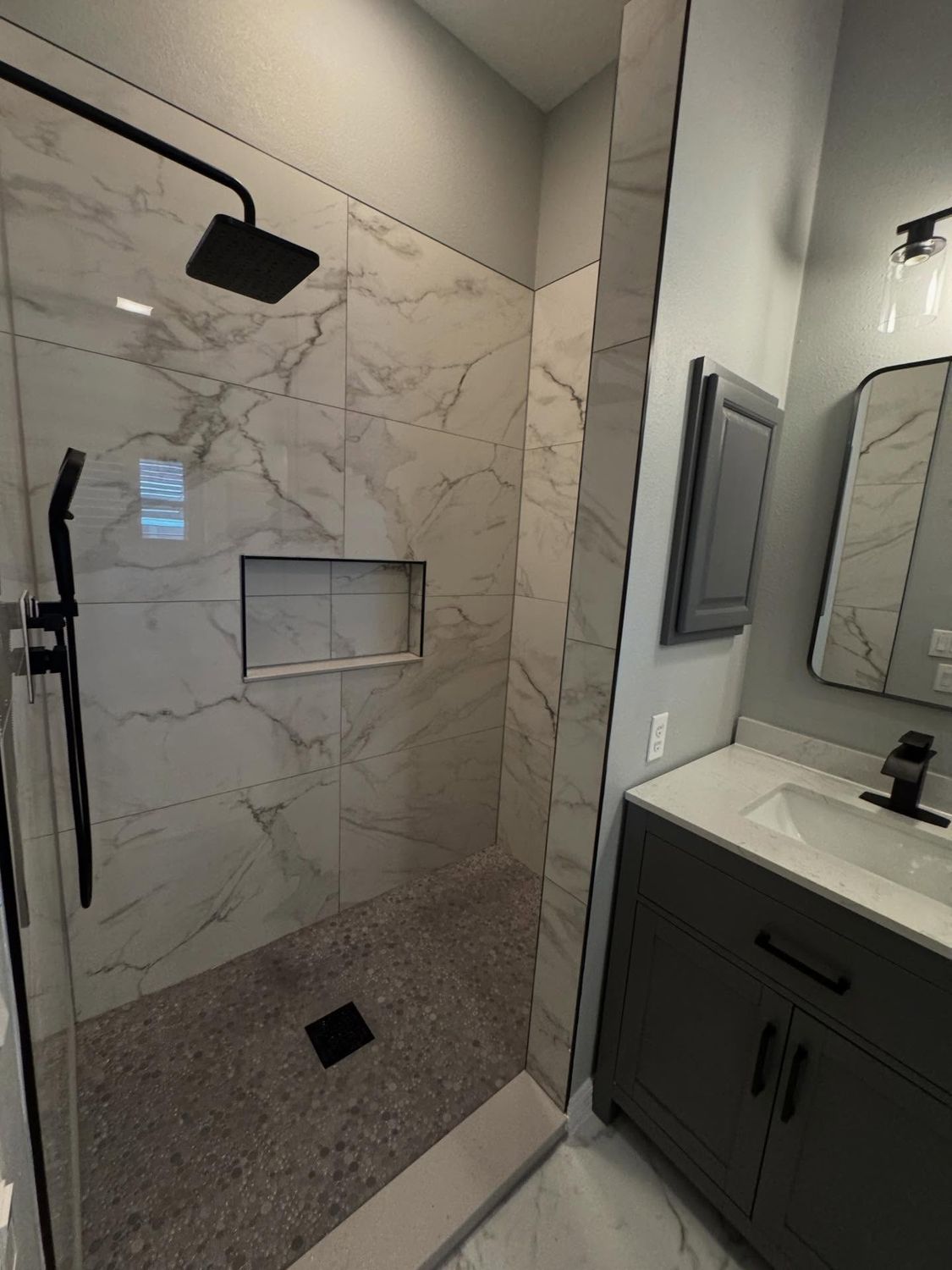 The same shower after remodeling, with full-height porcelain slab walls, a tile niche, pebble floor and matte black fixtures